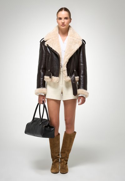 Jachetă bomber din piele brună cu guler din blană crem, cămașă albă, pantaloni scurți albi, cizme din suede brun deschis până la genunchi și o geantă neagră.