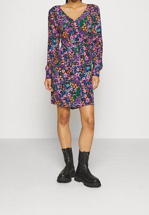 Femme portant une robe fleurie à manches longues, longueur genou, avec un col en V, associée à des bottines noires épaisses.