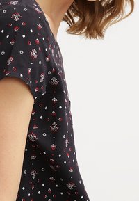 Tmavá látková blůza s krátkými rukávy, zdobená květinovým a geometrickým vzorem v červené, bílé a modré barvě, s volným střihem.