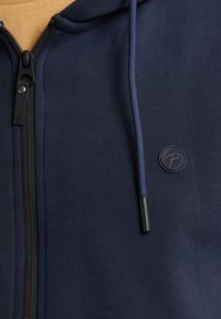 Navy zip-up hoodie made from soft fabric, featuring a black zipper, drawstring hood, and a round logo patch on the chest.