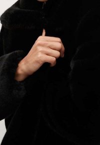 Czarna sztuczna futrzana kurtka z wysokim kołnierzem. Zbliżenie na rękę zapinającą przód. Miękka tekstura z gładkim wykończeniem i bez widocznych elementów metalowych.