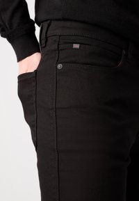 Jean en denim noir avec une coupe slim, des coutures visibles, une fermeture à bouton, et une étiquette de marque avec des accents rouges subtils près de la poche.