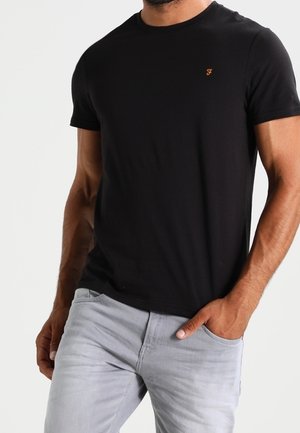 Mężczyzna w czarnej, krótkiej koszulce z małym pomarańczowym logo na piersi i jasnoszarych dżinsach, prawą ręką w kieszeni, na jednolitym tle.