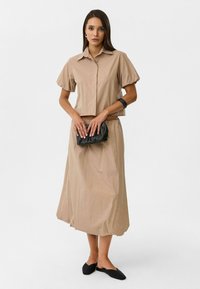 Beige korte mouwen blouse met knopen, gecombineerd met een bijpassende lange rok. Gelaagd weefsel, lichtgewicht; model houdt een zwarte clutch vast. Zwarte ballerina's.