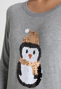 Grauer Pullover mit einem Pinguin-Design aus Pailletten. Der Pinguin trägt einen braunen Hut und einen Schal, der mit schwarzen, weißen und orangefarbenen Details verziert ist.