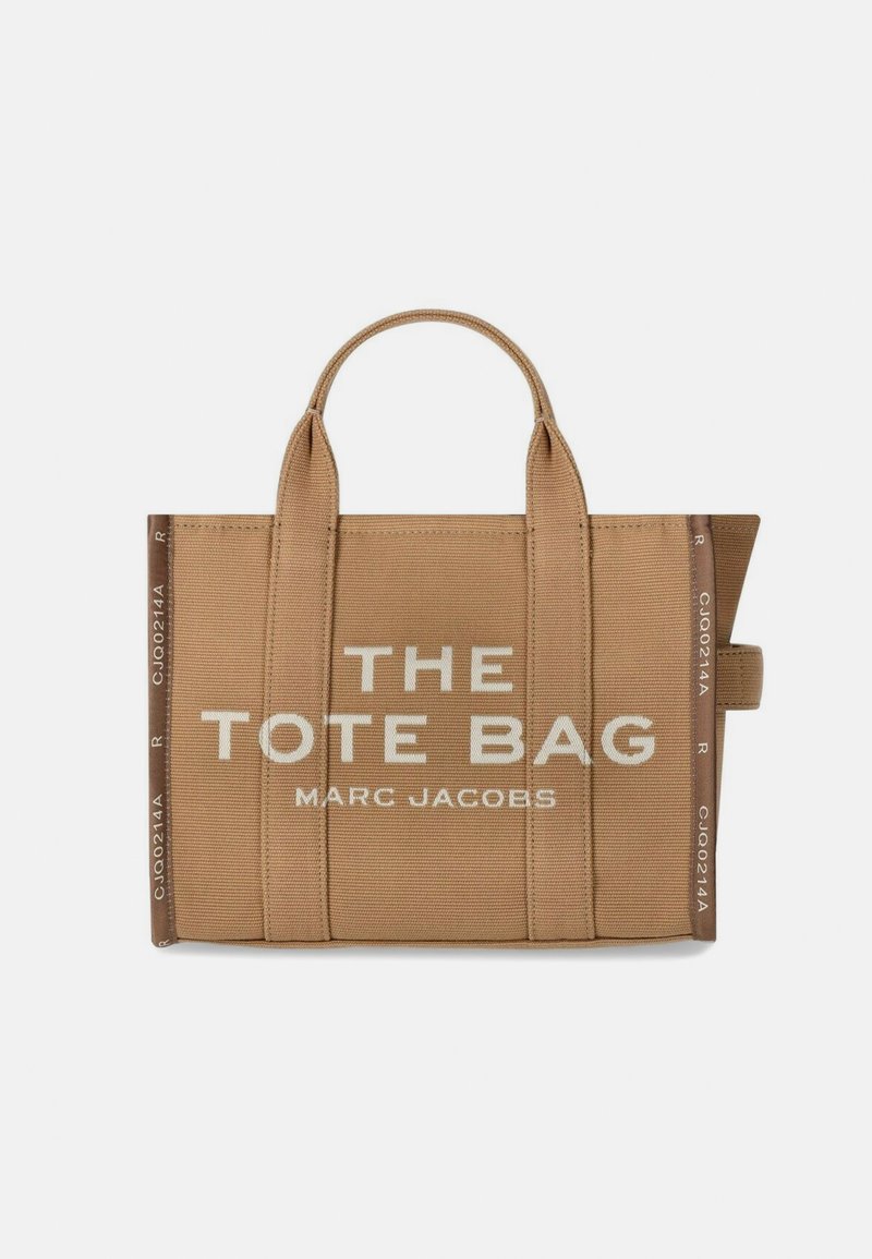 Tan spalvos kanvos totinis krepšys su dviem viršutiniais rankenomis. Ant krepšio yra baltu tekstu užrašai "THE TOTE BAG" ir "MARC JACOBS." Paprastas stačiakampio formos dizainas.