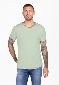 Camiseta de algodón verde claro con cuello redondo y mangas cortas, combinada con jeans azules desgastados. Presenta tatuajes visibles en el brazo.