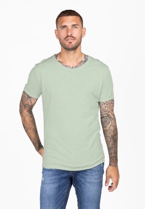 Lichtgroene katoenen t-shirt met een ronde halslijn en korte mouwen, gecombineerd met versleten blauwe jeans. Heeft zichtbare tatoeages op de arm.