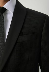 Musta puku takki, jossa on teksturoitu kuvio, satiinikaulus ja pieni rintatasku. Yhdistetty mustan solmion kanssa, jossa on hienovaraisia pisteitä.