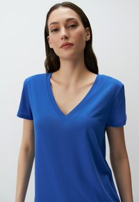 Niebieska koszulka z dekoltem w serek, krótki rękaw, wykonana z miękkiego materiału, gładka w dotyku, o luźnym kroju, z minimalnymi detalami szycia.