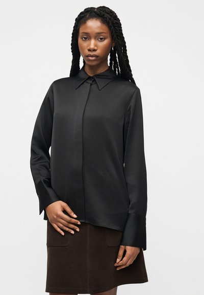 Γυναίκα με μακριά πλεγμένα μαλλιά, φοράει μαύρο πουκάμισο με κουμπιά και επιμηκυμένες μανσέτες και σκούρα καφέ φούστα, στέκεται μπροστά από ένα απλό φόντο.