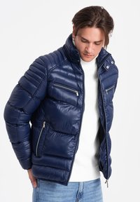 Granatowa kurtka puchowa z pikowanym wzorem, wysokim kołnierzem i kieszeniami na zamek. Wykonana z błyszczącego materiału o dopasowanym kroju.