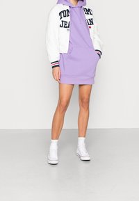 Lyslilla hættetrøje-kjole med frontlomme, parret med en hvid quiltet bomberjakke med marineblå og røde accenter. Hvide sneakers fuldender looket.