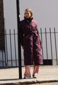 Gilet lungo imbottito di un profondo bordeaux con vita elasticizzata e texture a stelle. Abbinato a pantaloni a gamba larga rosa pallido e sneakers.