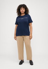 Jūras spēku zila kokvilnas T-kreklā ar "TOMMY HILFIGER" logo, apvienots ar beidža straight-leg biksēm un melnām mokasīnām. Gluda tekstūra, ikdienišķa piegriezuma.