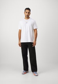 Hvit bomulls t-skjorte med en liten rød logo, sammen med svarte teksturerte bukser og flerfargede joggesko. Rett snitt-design, minimale mønstre.