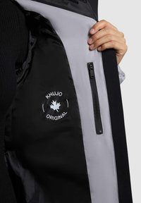 Giubbotto nero con fodera grigia, dotato di tasca con zip e un logo che recita "Khujo Original" con un design a foglia d'acero.