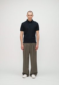 Svart pikétröja med korta ärmar, kombinerad med lösa, ljus olivgröna byxor. Modellen bär vita sneakers och står mot en neutral bakgrund.