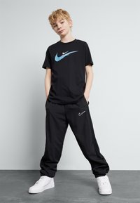 T-shirt noir avec logo Nike bleu et blanc, pantalon noir avec accent blanc Nike et baskets blanches. Design simple et sportif avec une coupe décontractée.