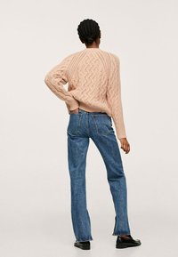 Personne aux cheveux tressés portant un pull en maille torsadée rose clair, un jean bleu droit et des mocassins noirs, debout avec une main dans la poche.