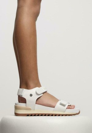 Sandalias blancas con un upper texturizado, correa ajustable y una suela gruesa y estriada. El diseño cuenta con un acento metálico circular.