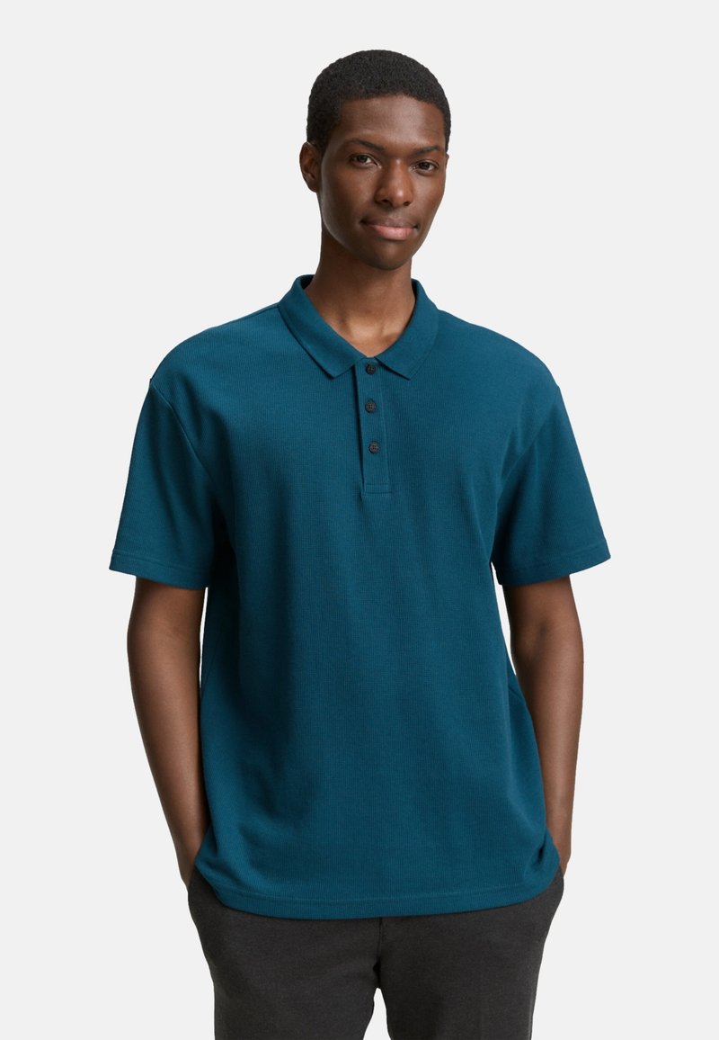 Tengerés kék pamut póló rövid ujjakkal, gallérral és három fekete gombbal a nyaknál. Sima textúra és laza fazon.