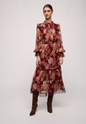 Lang maroon jurk met bloemenprint, gerimpelde kraag en mouwen, kantafwerking aan de zoom en een stoffen riem. Gekleed met bruine kniehoge laarzen.
