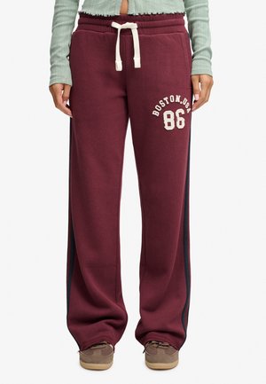 Person iført bordeaux sweatpants med hvid snøre og teksten "Boston, USA 86", kombineret med brune sneakers og en lysegrøn top.