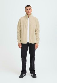 Beige fleece jack met een hoge kraag, lange mouwen en rits. Draagt over een wit shirt. Zwarte broek en schoenen met witte strepen.