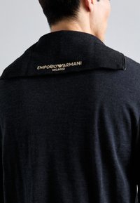 Uomo che indossa un maglione nero Emporio Armani Milano, visibile da dietro, con il logo sul colletto ripiegato su uno sfondo semplice.