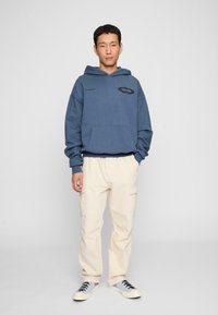 Felpa in cotone blu con tasca frontale, caratterizzata da una grafica nera e del testo. Abbinata a pantaloni cargo crema e sneakers con dettagli blu.