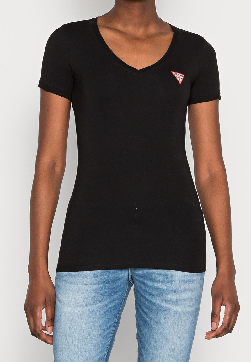 T-shirt noir à manches courtes en V avec petit logo triangulaire rouge et blanc sur la poitrine gauche, porté avec un jean bleu clair.
