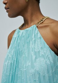 Une robe plissée bleu clair présente un motif floral texturé. L'encolure est ornée d'un détail de chaîne en or.