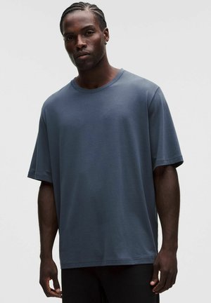 Hombre con el cabello trenzado que lleva una camiseta de manga corta suelta de color azul oscuro, de pie frente a un fondo liso y claro.