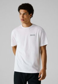 Camiseta de algodón blanca de manga corta, con un pequeño logo negro de "Red Bull Racing" en el lado izquierdo del pecho. Diseño de cuello redondo estándar.
