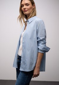 Femme portant une chemise à rayures verticales bleu clair boutonnée par-dessus un haut blanc, manches retroussées, et un jean bleu foncé, debout devant un fond uni.