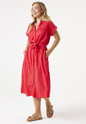 Vestido rojo de longitud a la rodilla con mangas cortas bordadas, un frente abotonado y una cintura ajustada. Llevado con sandalias de tacón bajo color beige.