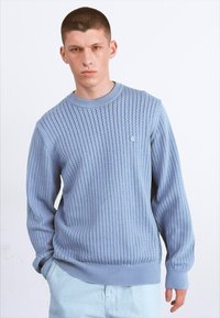 Suéter de punto azul claro con un patrón texturizado, cuello y puños acanalados, y un pequeño logo de acento en el lado izquierdo del pecho.