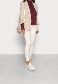 Femme portant un blazer beige, un col roulé bordeaux, un pantalon skinny blanc, des mocassins à plateforme beiges et un sac à épaule beige texturé.