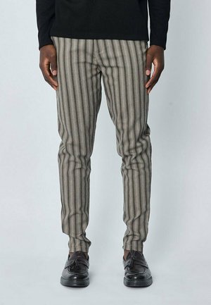 Personne portant un pantalon gris à rayures verticales et des mocassins noirs à pampilles, debout devant un fond clair uni.