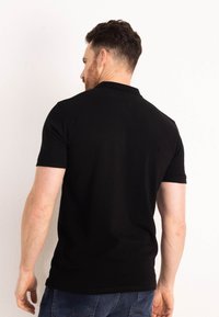 Černé bavlněné polo tričko s krátkým rukávem, žebrovaným límcem a postranními rozparky. Spodní lem je mírně zakřivený, což zajišťuje uvolněný střih.