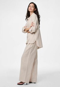 Outfit due pezzi di un beige chiaro e strutturato, caratterizzato da una camicia button-up ampia e pantaloni a gamba larga, abbinato a sandali neri con tacco.