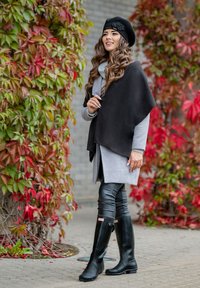 Poncho noir sur un pull gris, associé à des leggings noirs ajustés et des bottes en caoutchouc noires. Stylé avec un béret noir et un accent floral.