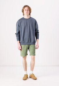 Sivý sveter s okrúhlym výstrihom v kombinácii s olivovo zelenými šortkami. model má na sebe béžové tenisky a biele ponožky, stojí na neutrálnom pozadí.