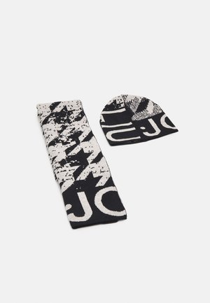 Schwarz-weiß gemusterter Strickschal und passende Mütze mit abstrakten Formen und großen Buchstaben, flach auf einer weißen Oberfläche ausgelegt.