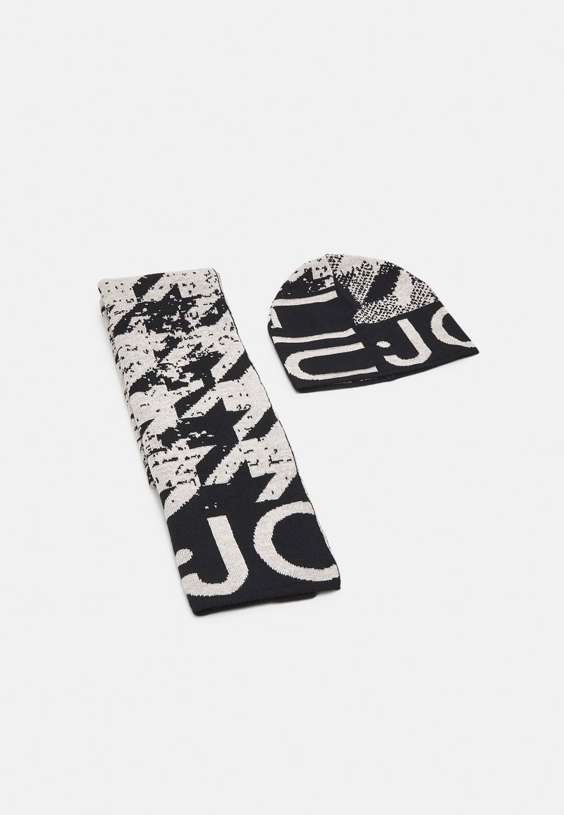 Écharpe tricotée à motifs noir et blanc et bonnet assorti avec des formes abstraites et de grandes lettres posés à plat sur une surface blanche.