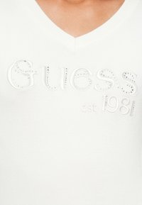 Maglione bianco a V con logo "Guess" in rilievo e "est. 1981", con dettagli e texture accentuati da strass.