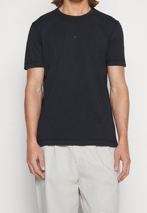 Man wearing a plain black short-sleeve t-shirt with small "BOSS" logo on chest and light beige pants against a white background.