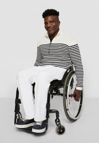 Gestreepte witte en marineblauwe trui met een halve rits, gecombineerd met witte broek en marineblauwe sneakers. Persoon zit in een rolstoel en glimlacht.