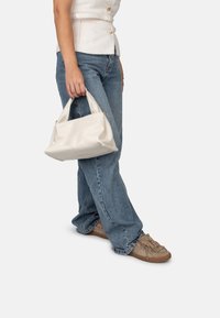 Borsa a tracolla sintetica bianca tenuta in una mano, con un design morbido e strutturato e un solo manico. Indossata con jeans blu e scarpe beige.